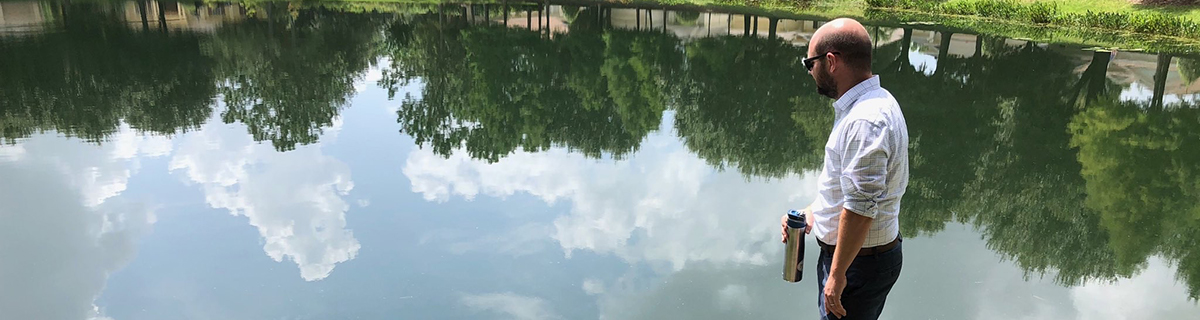 Man standing over very calm pond reflecting the sky and trees