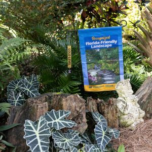 A gold-level FFL flag in a home landscape surrounded by green, shade-loving foliage.